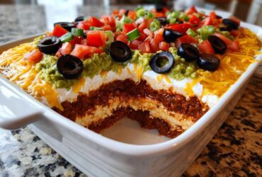 A white dish showing a cross-section of a delicious 7 layer dip topped with guacamole, tomatoes, olives, and cheese.
