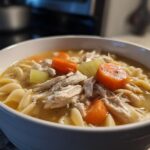 Close-up of a white bowl filled with amazing 1-pot chicken noodle soup, featuring shredded chicken, egg noodles, carrots, and celery.