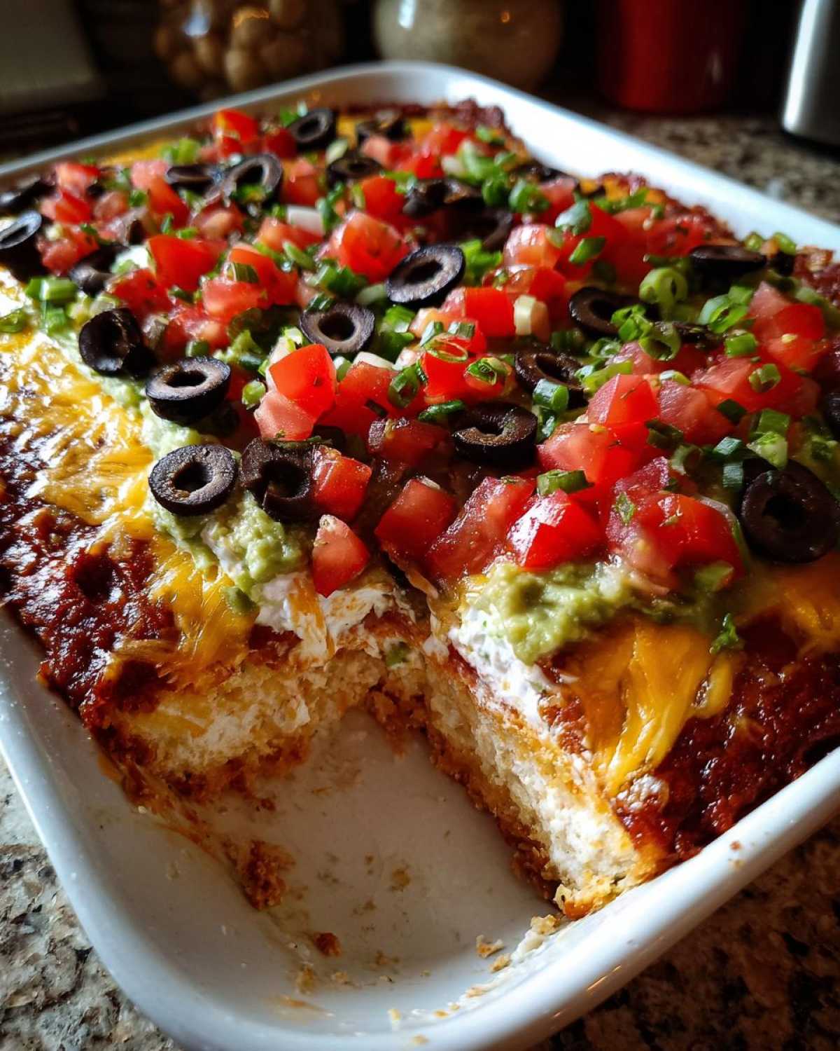 Close-up of a baked 7 layer dip in a white dish, showing layers of cheese, sour cream, guacamole, tomatoes, and olives.