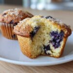 Close-up of a freshly baked blueberry muffin cut in half showing moist interior and large blueberries, part of a batch of blueberry muffins.
