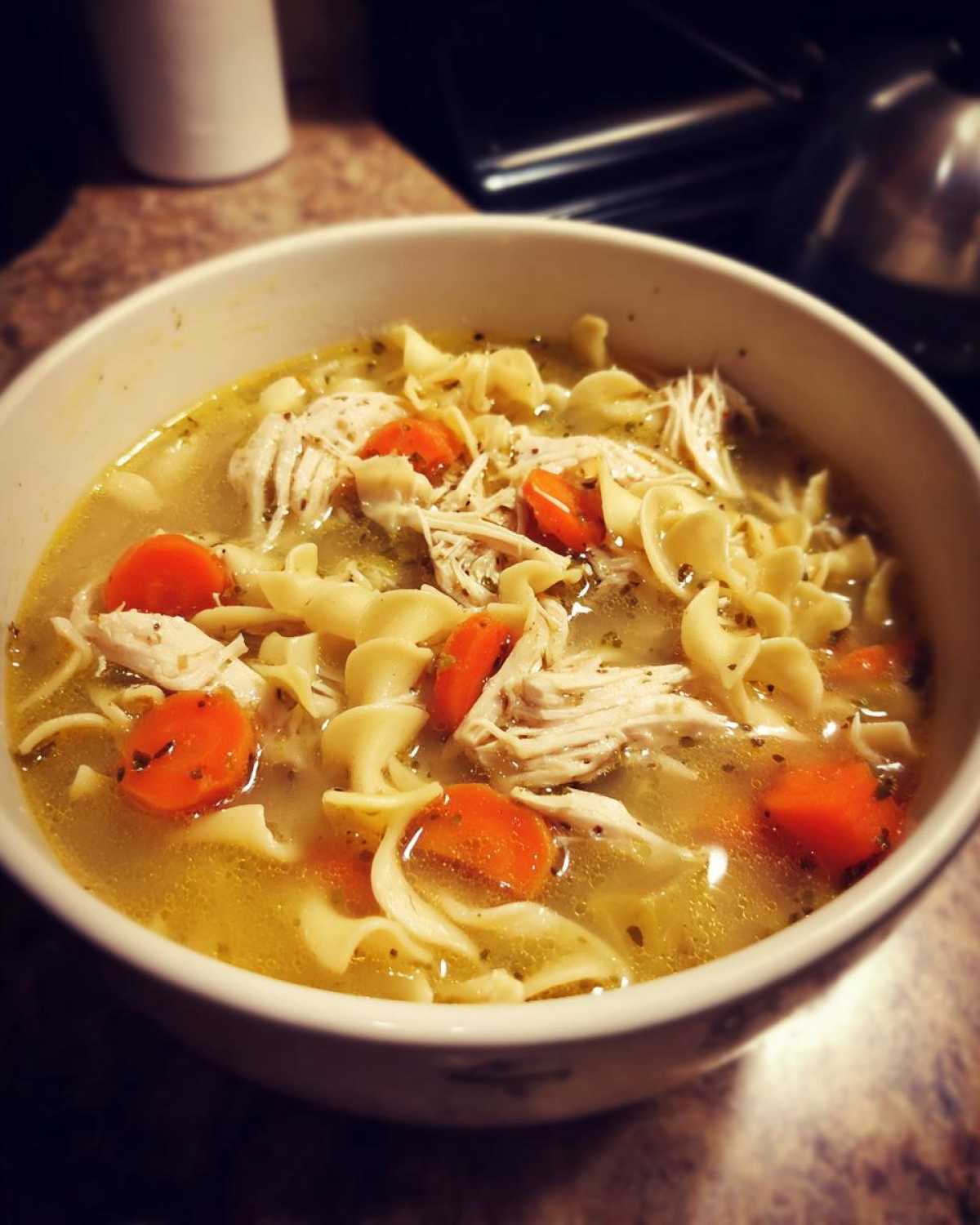 Close-up of a white bowl filled with homemade chicken noodle soup, featuring shredded chicken, egg noodles, and bright orange carrots.