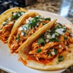 Close-up of three soft corn tortillas filled with shredded chicken for amazing chicken tacos, topped with diced white onion and cilantro.