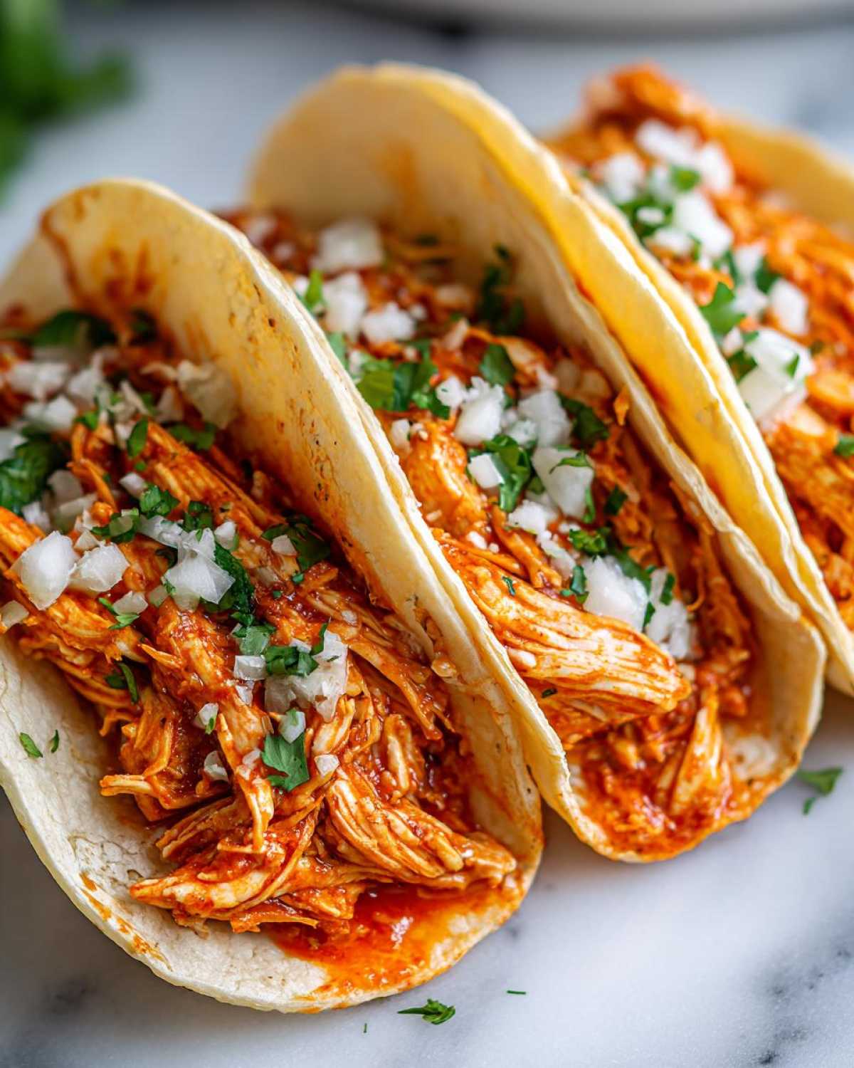 Close-up of three soft tortilla chicken tacos filled with saucy shredded chicken, topped with diced white onion and cilantro.