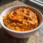 Close-up of a thick, vibrant orange chili cheese dip with visible red chili flecks, served in a white bowl.