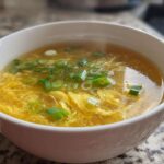 A close-up of a white bowl filled with steaming egg drop soup, featuring delicate yellow egg ribbons and green scallions.