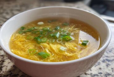 A close-up of a white bowl filled with steaming egg drop soup, featuring delicate yellow egg ribbons and green scallions.