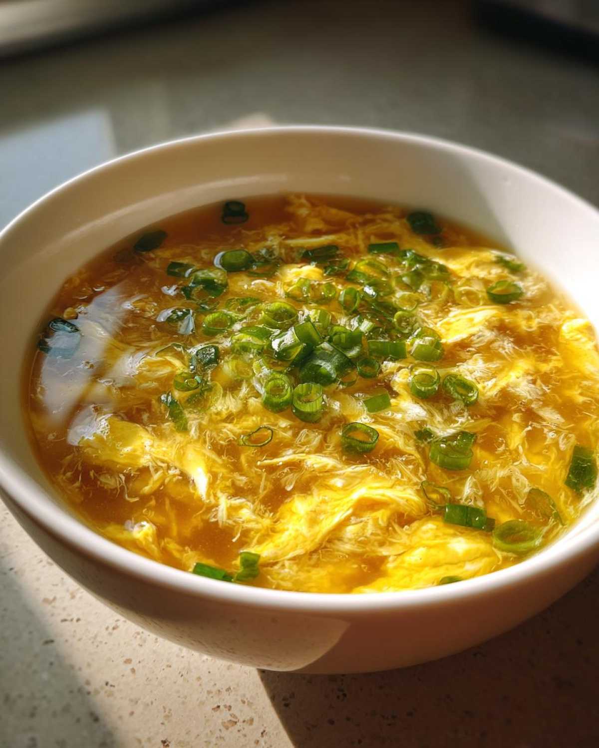 A close-up of a white bowl filled with hot egg drop soup, featuring wispy yellow egg ribbons and topped with fresh green scallions.