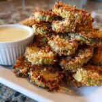 A pile of golden brown, crispy Fried Pickles served on a white plate next to a small bowl of creamy dipping sauce.