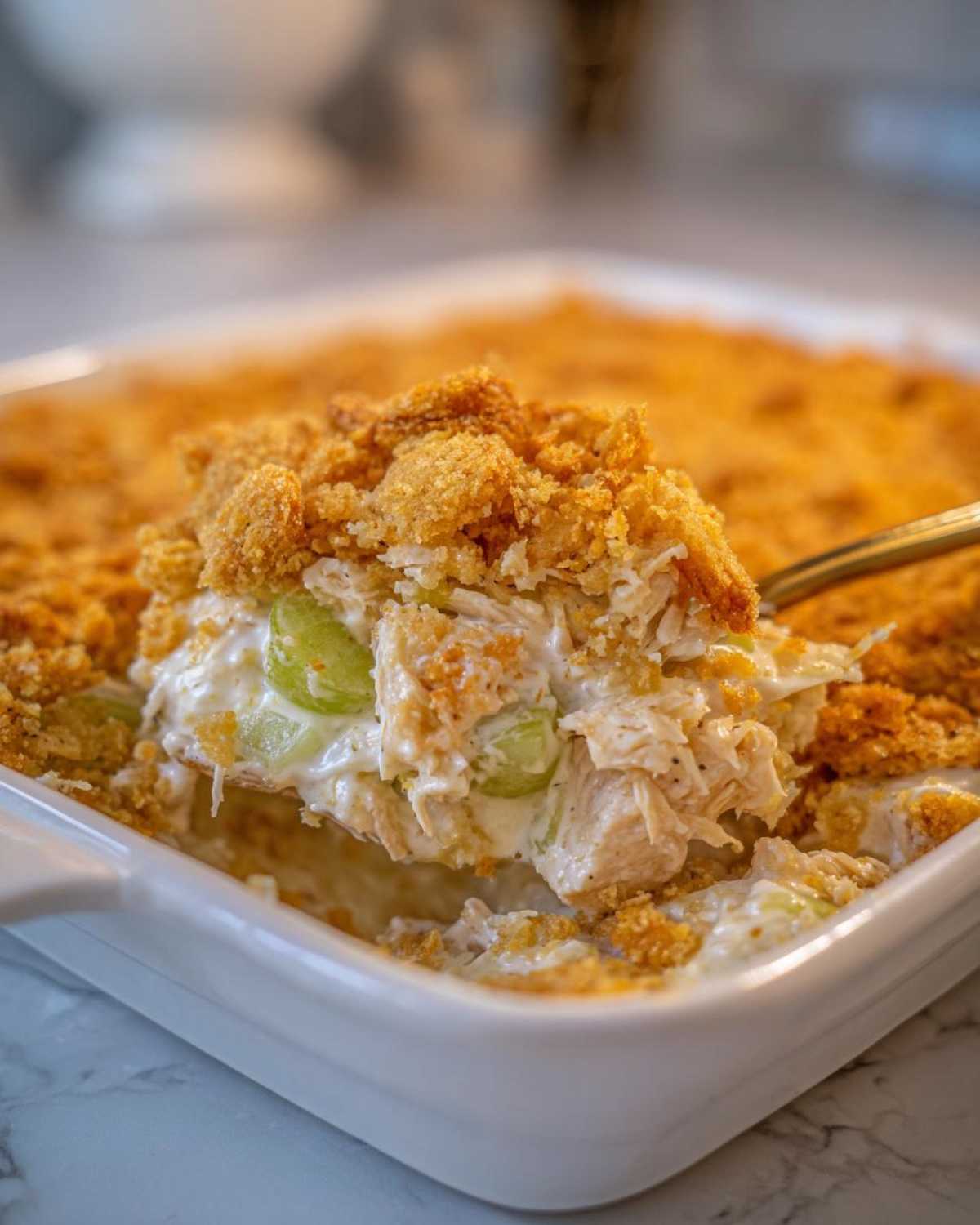 A spoonful of creamy hot chicken salad with celery and a crunchy topping being lifted from a white baking dish.