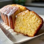 Close-up of a moist lemon pound cake loaf with a thick white glaze dripping down the sides, one slice cut.