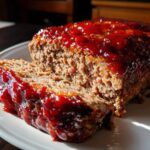 Close-up of a juicy meatloaf with a thick, shiny red glaze, showing one slice cut and resting against the main loaf.
