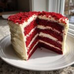 A stunning, tall slice cut from a red velvet cake showing four layers of deep red cake separated by thick white cream cheese frosting.