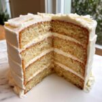 A tall, four-layer vanilla cake recipe cut to show the moist crumb and thick white frosting layers.