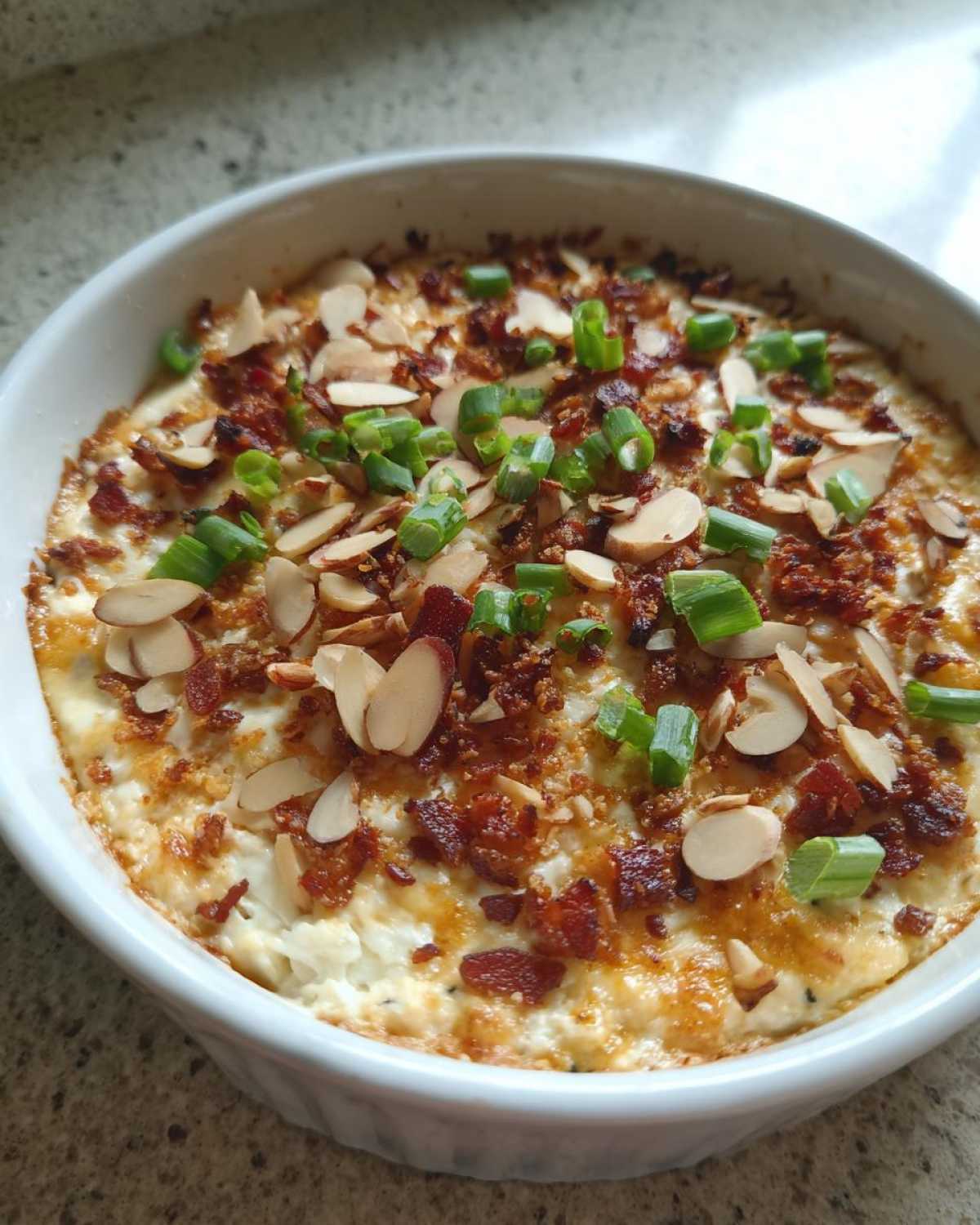 Close-up of hot, baked neiman marcus dip topped with sliced almonds, bacon bits, and chopped green onions.