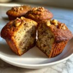 A banana protein muffin cut in half showing the moist interior texture, topped with walnuts.