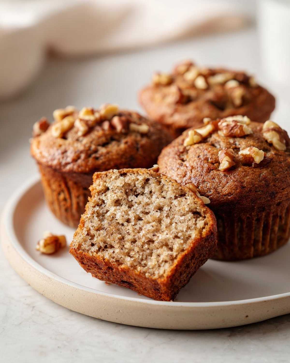 Close-up of a banana protein muffin cut in half showing the moist crumb, served with whole muffins topped with walnuts.