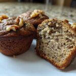 Three banana protein muffins on a white plate, one cut in half showing the moist interior and topped with walnuts.