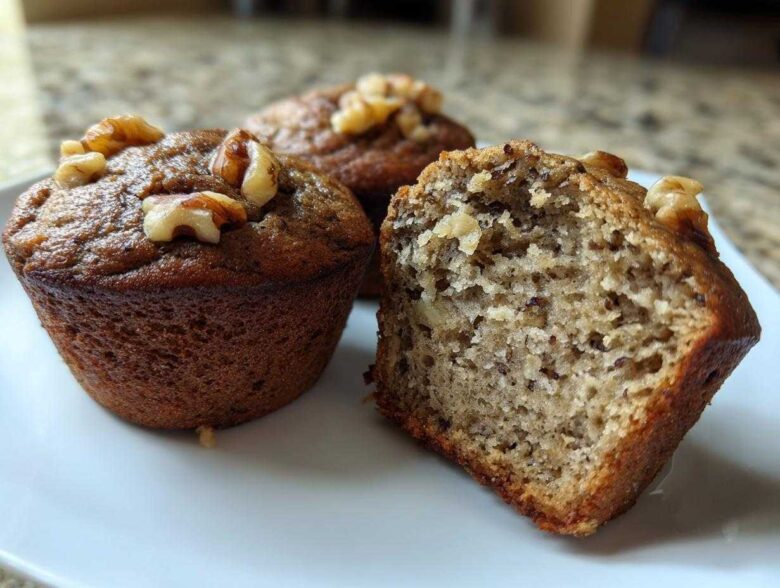 Three banana protein muffins on a white plate, one cut in half showing the moist interior and topped with walnuts.