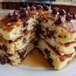 A thick stack of fluffy chocolate chip pancakes cut in half, showing melted chocolate inside and topped with chocolate chips.