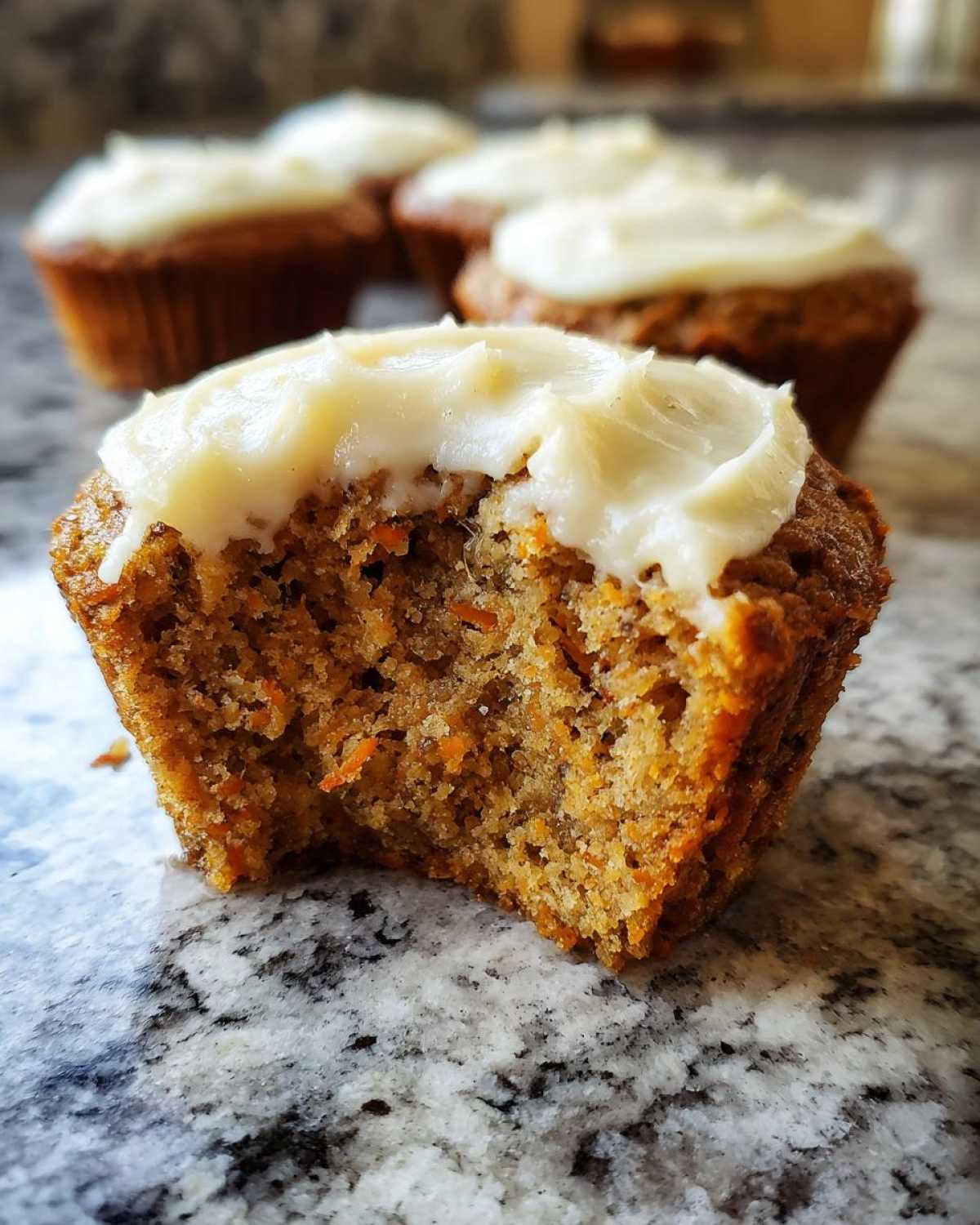 Close-up of a carrot cake muffin with a bite taken out, showing moist texture and cream cheese frosting.