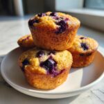 A stack of four golden-brown blueberry protein muffins sitting on a white plate in bright sunlight.