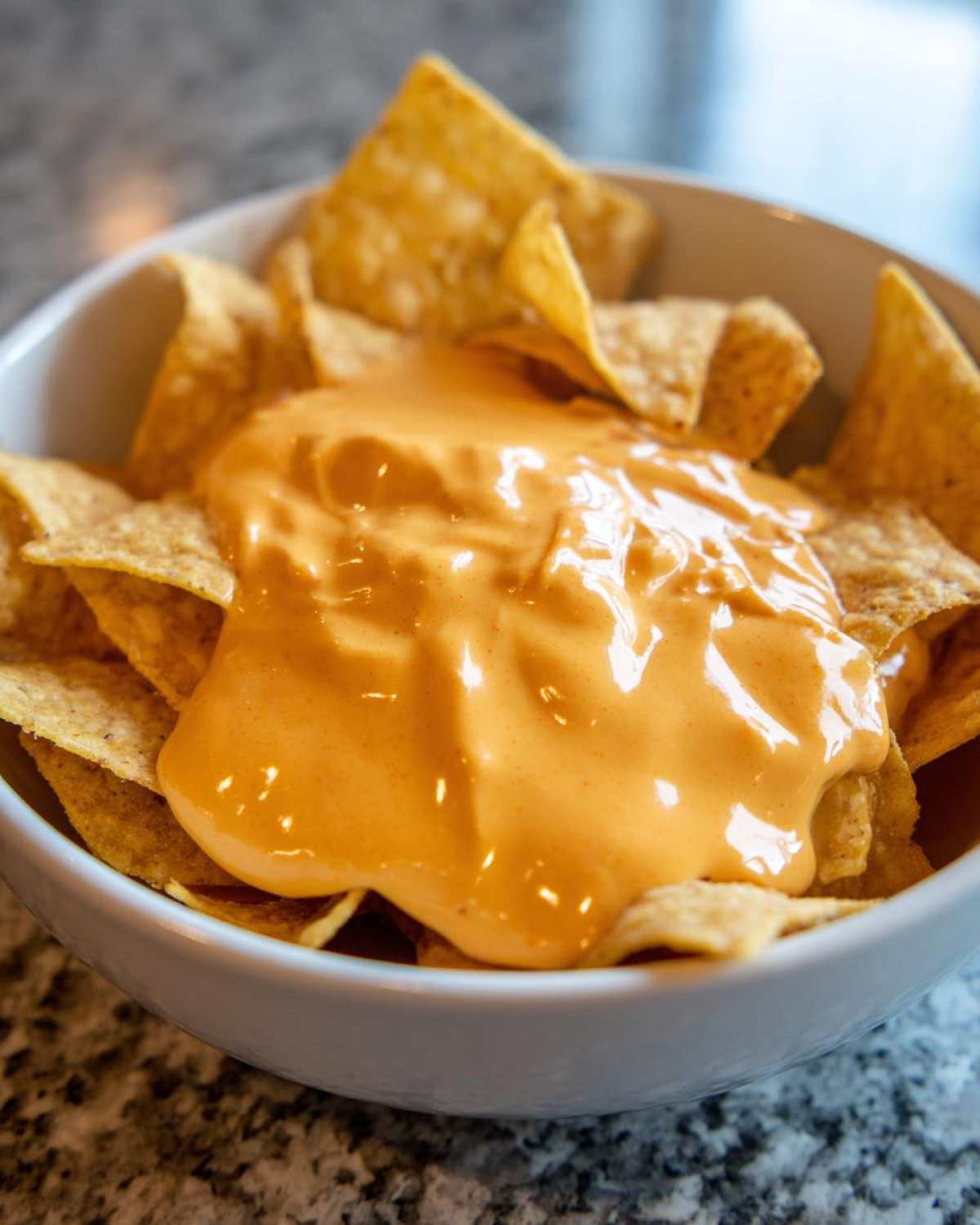 Close-up of tortilla chips smothered in creamy, bright orange nacho cheese sauce in a white bowl.
