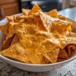 A close-up of tortilla chips generously smothered in creamy, bright orange nacho cheese sauce in a white bowl.