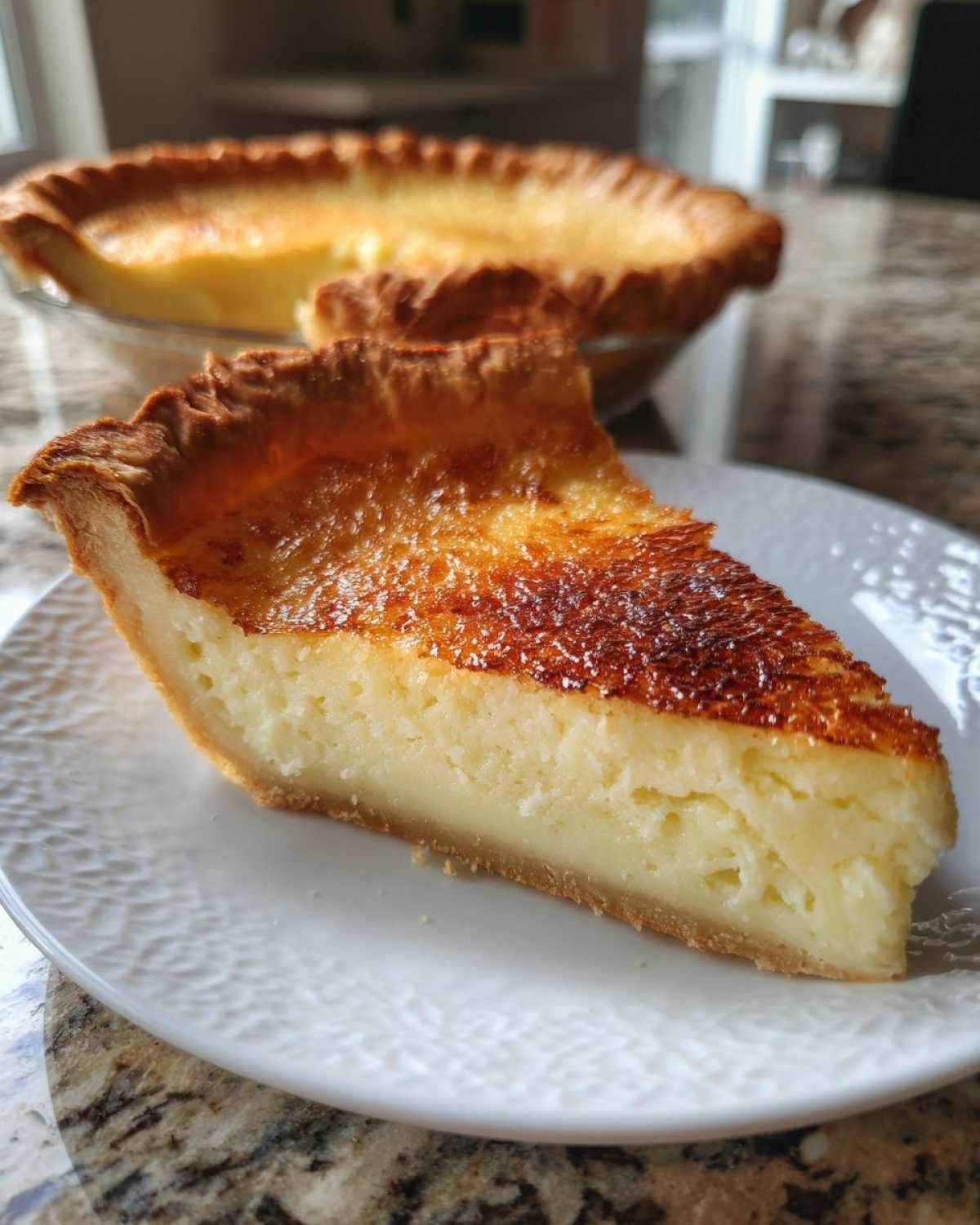 A thick slice of creamy buttermilk pie with a deeply caramelized top crust sits on a white plate.