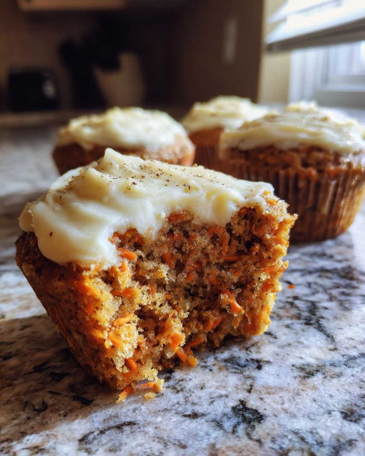 A close-up view of a carrot cake muffin with a bite taken out, revealing shredded carrots, topped with cream cheese frosting and spices.
