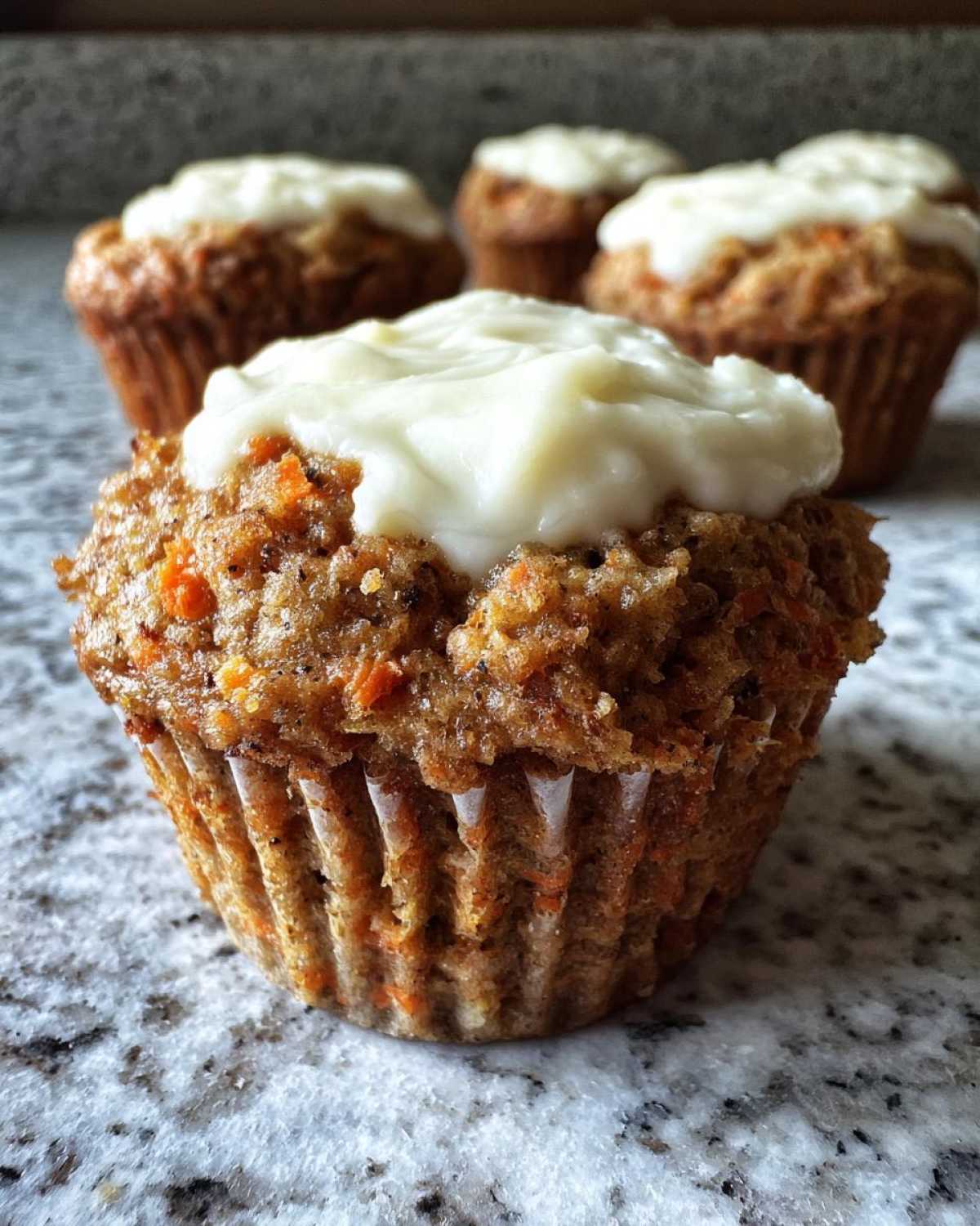 A close-up of a single, moist carrot cake muffin topped with thick cream cheese frosting, with more carrot cake muffins blurred in the background.