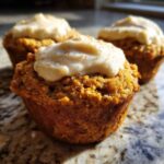 A close-up of a delicious carrot cake muffin topped with thick cream cheese frosting and a dusting of spice.
