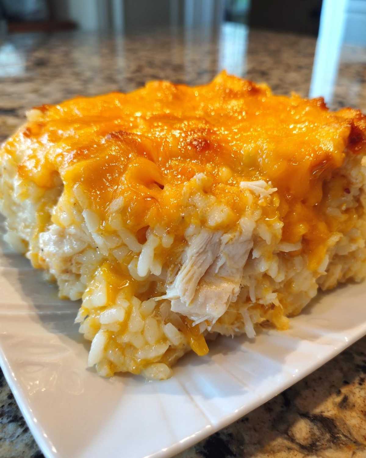 A close-up of a square serving of cheesy chicken and rice casserole topped with melted cheddar cheese.