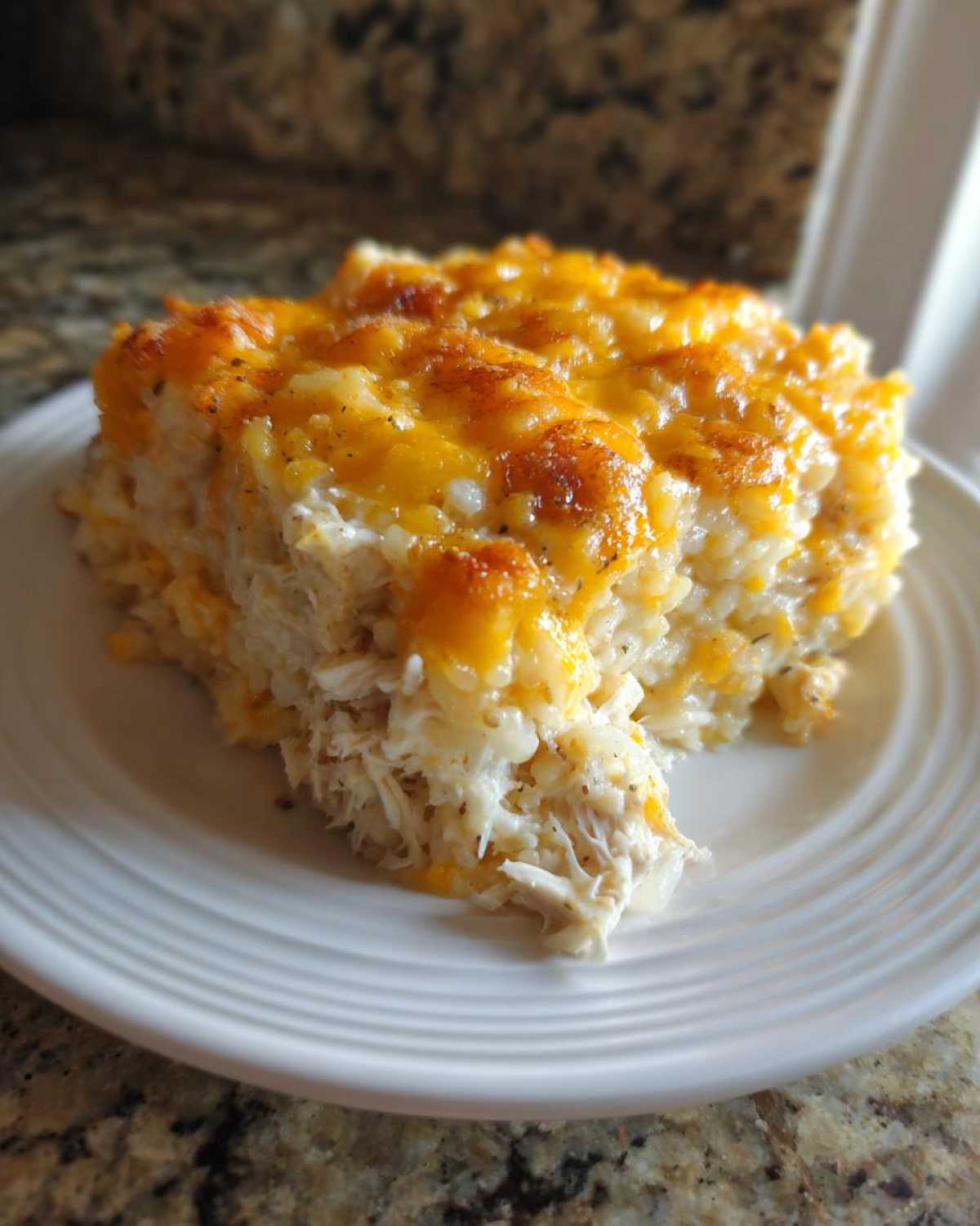 A square serving of cheesy chicken and rice casserole with a golden melted cheese topping on a white plate.