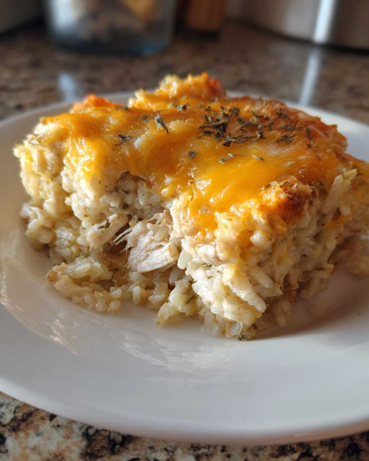 A close-up serving of cheesy chicken and rice casserole topped with melted cheddar and dried herbs.