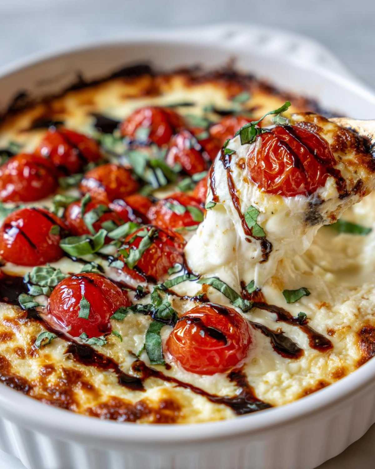 A spoonful of hot, cheesy bruschetta dip is pulled from the baking dish, showing melted cheese strings, roasted tomatoes, and balsamic glaze.