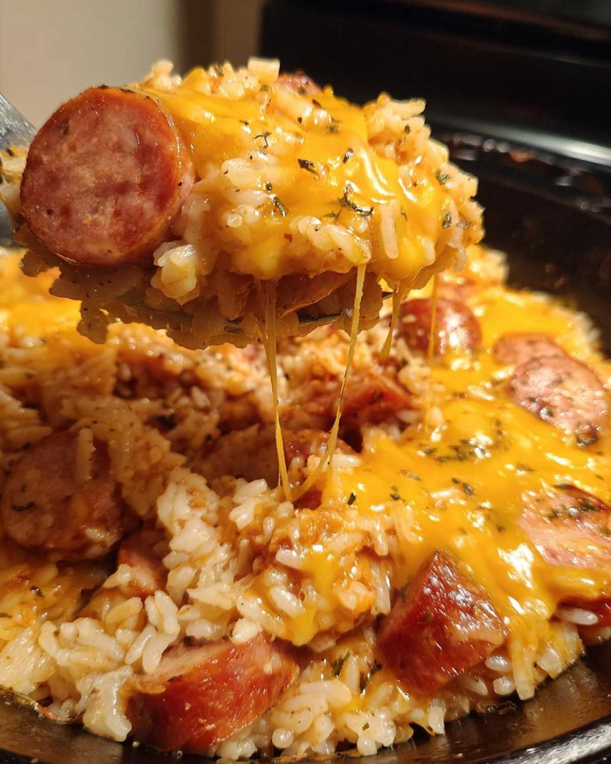 Close-up of a spoonful of cheesy sausage and rice, showing melted cheese stretching between the spoon and the bowl.