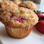 A close-up of a freshly baked strawberry muffin topped with cinnamon sugar, next to whole fresh strawberries.