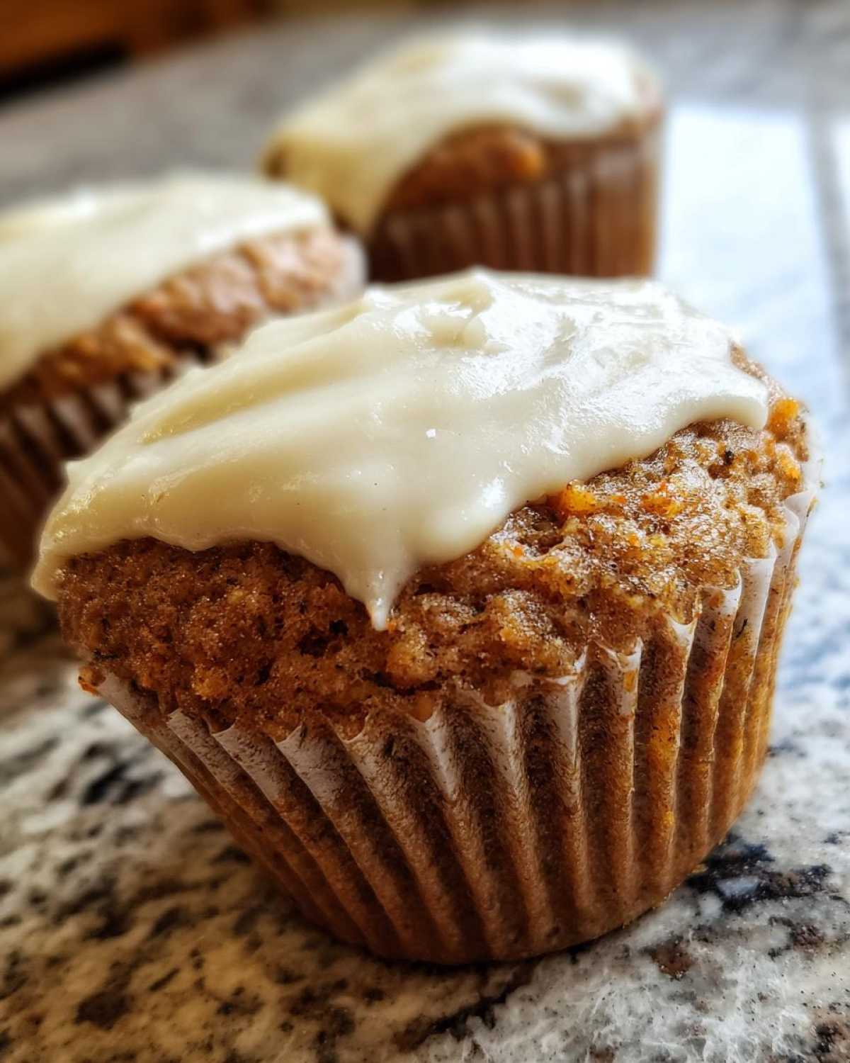 A close-up of a moist carrot cake muffin topped with thick, creamy white frosting.