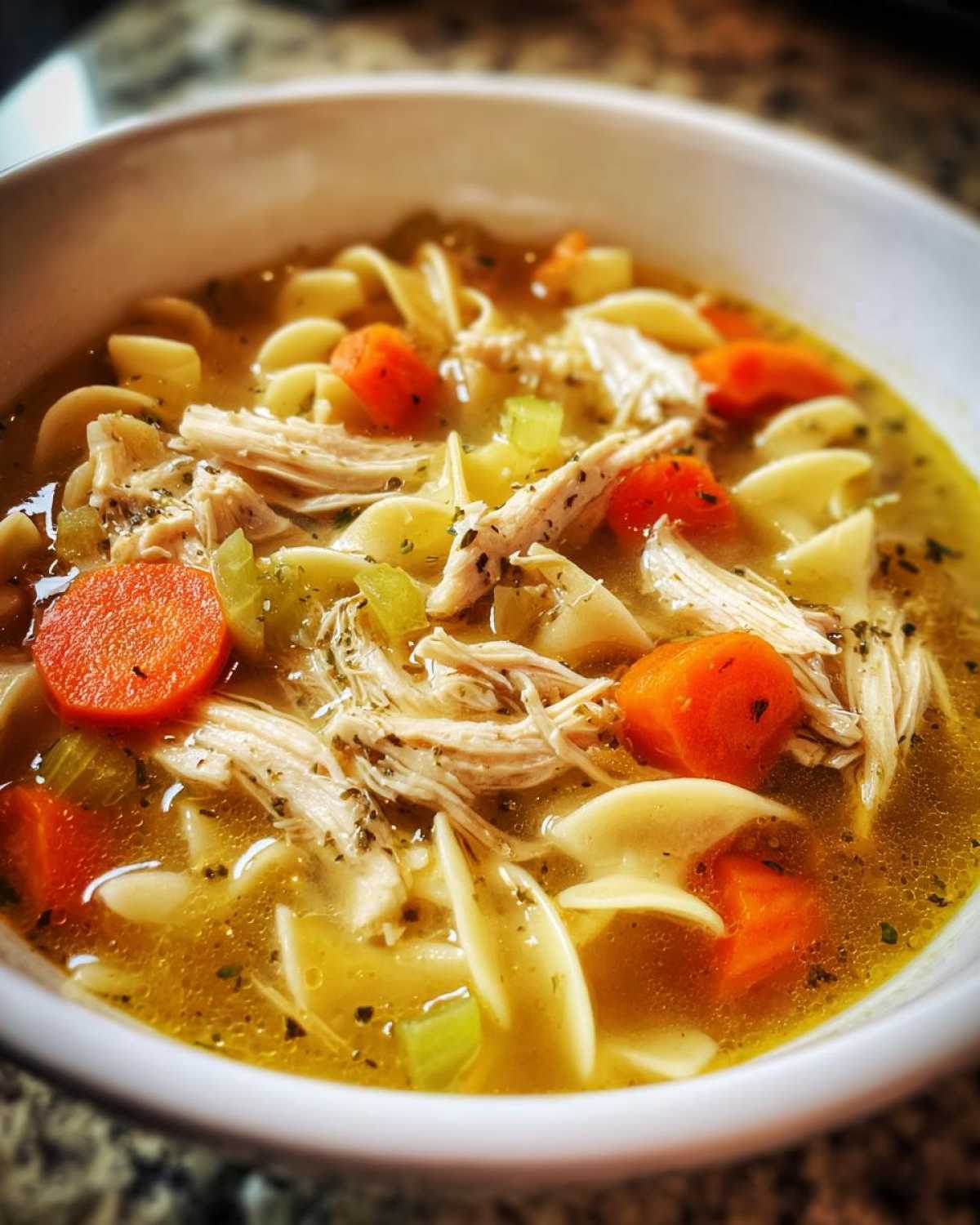 Close-up of a white bowl filled with homemade chicken noodle soup, featuring shredded chicken, egg noodles, carrots, and celery.