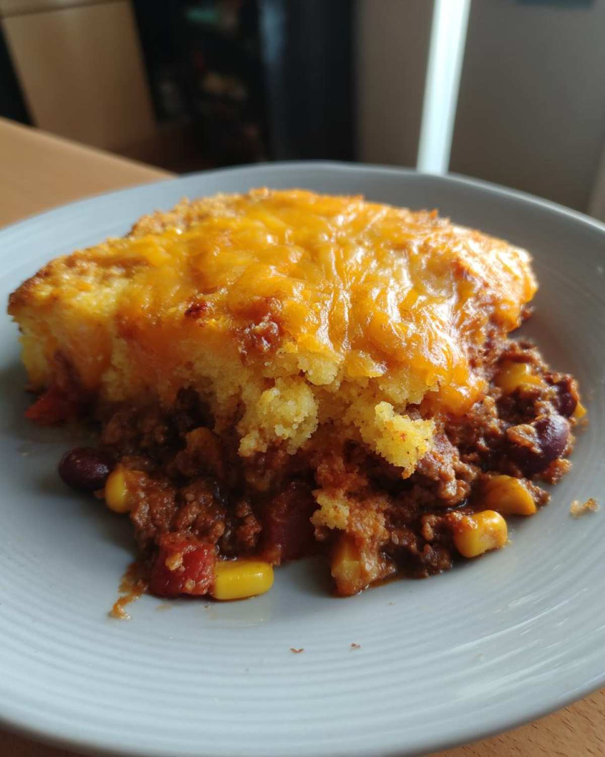 A serving of cowboy cornbread casserole topped with melted cheddar cheese resting on a bed of chili with corn and beans.