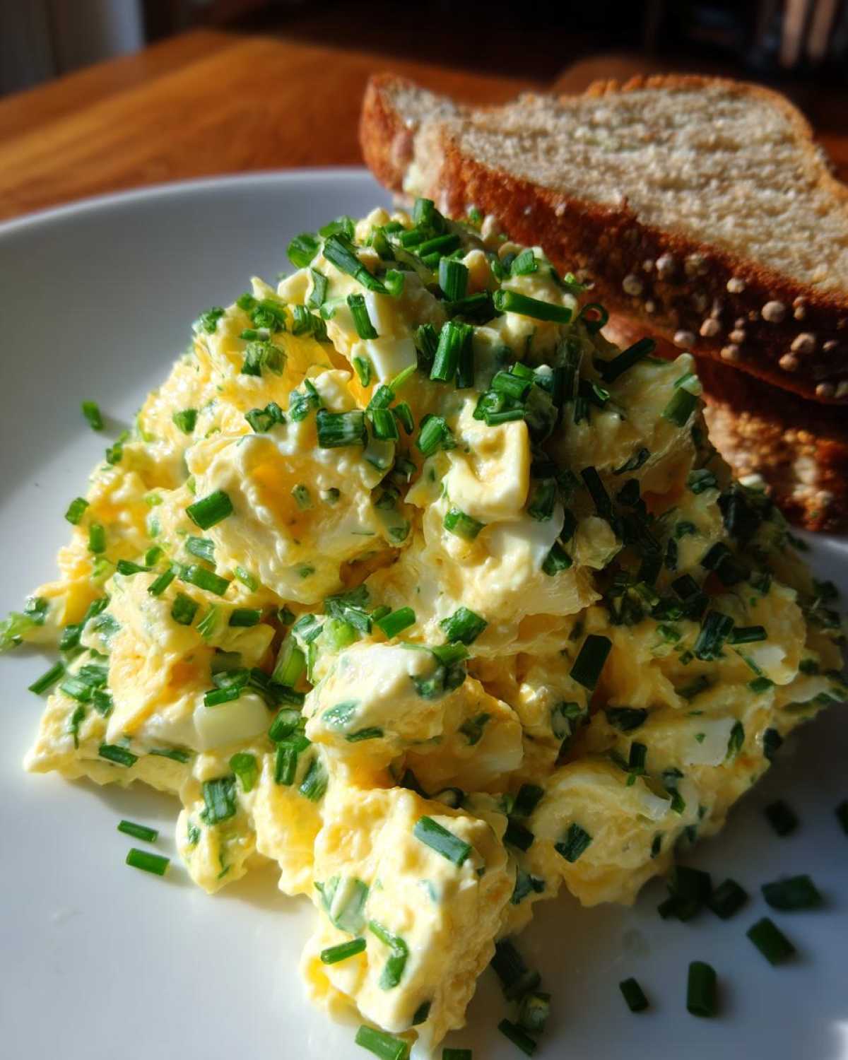 A mound of creamy egg salad recipe generously topped with bright green chopped chives, served next to slices of whole-grain bread.