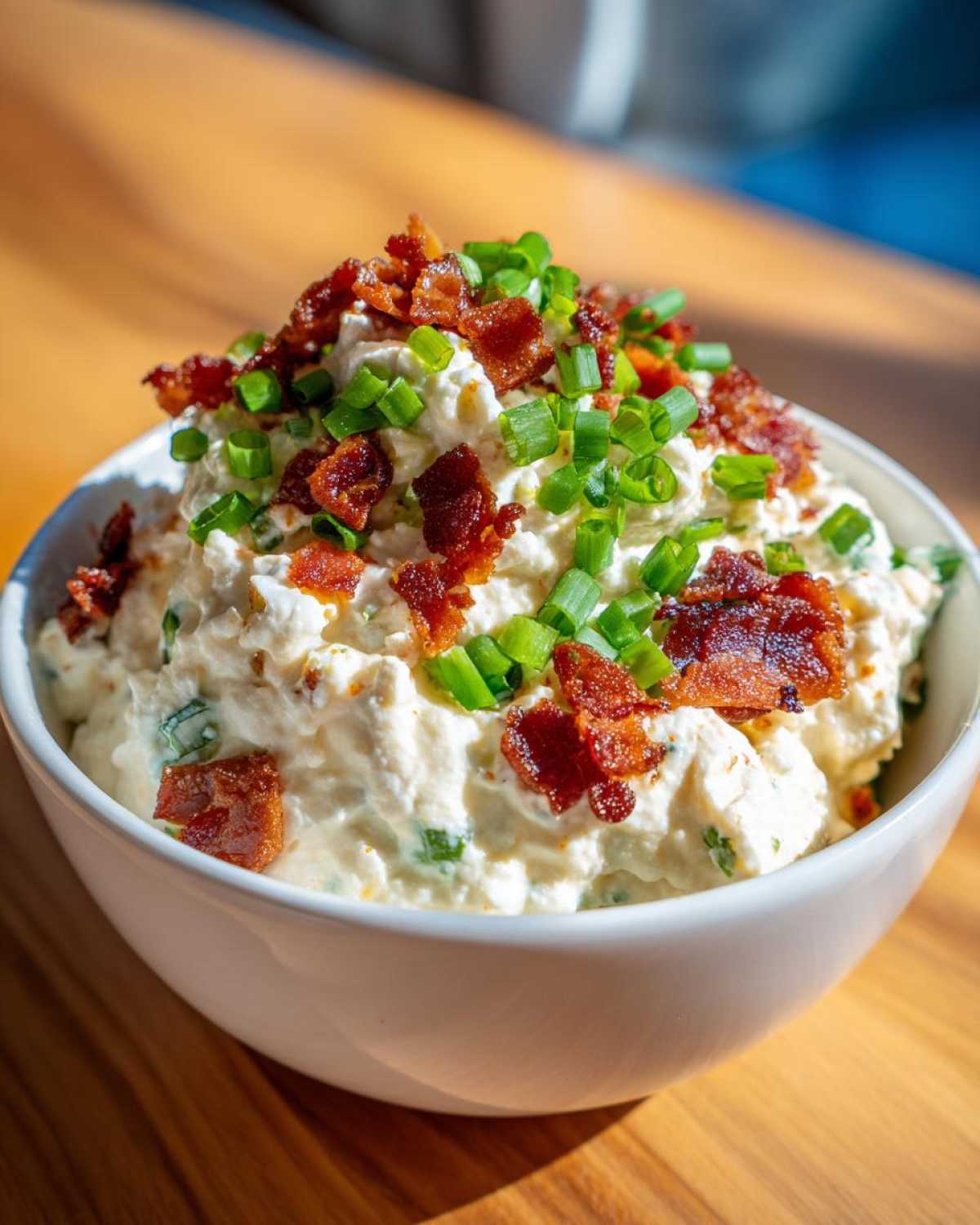 A close-up of creamy Million Dollar Dip served in a white bowl, generously topped with crispy bacon bits and fresh green chives.