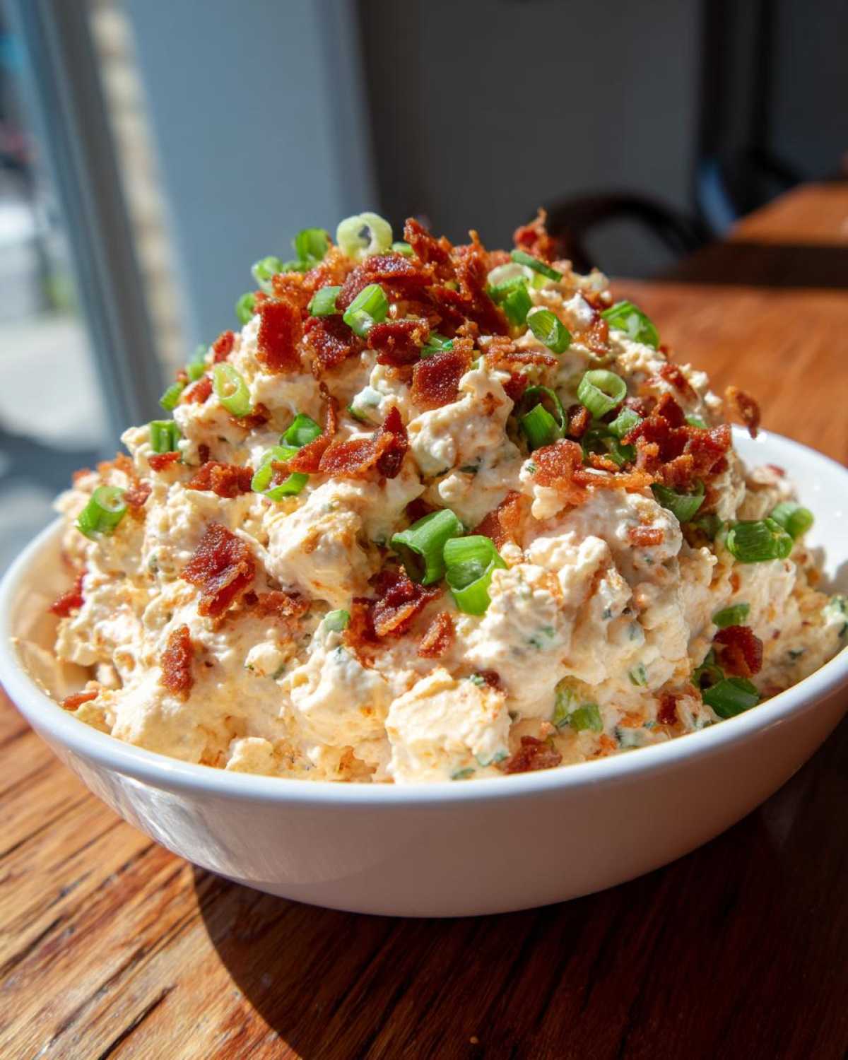 A creamy, orange-tinted million dollar dip piled high in a white bowl, topped with crispy bacon bits and green onions.