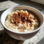 A close-up of creamy rice pudding served in a white bowl, generously sprinkled with ground cinnamon.