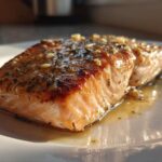A perfectly cooked fillet of air fryer salmon, glazed with herbs and garlic, resting in its juices on a white plate.
