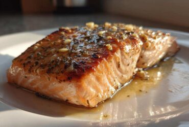 A perfectly cooked fillet of air fryer salmon, glazed with herbs and garlic, resting in its juices on a white plate.