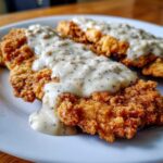 Close-up of two pieces of crispy chicken fried steak smothered in creamy, black pepper-flecked gravy.
