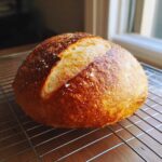 A beautifully baked, round loaf of crusty Italian bread with a golden-brown crust cooling on a wire rack.