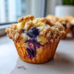 A close-up, featured image of a golden brown blueberry muffin topped with a generous layer of sugary streusel topping.