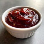 A close-up of thick, glossy, dark red homemade bbq sauce served in a small white ramekin.